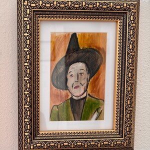 May include: A framed watercolor painting of a person wearing a black witch hat and a green jacket. The person has a white face with dark eyes and a dark mouth. The background is a light brown color.