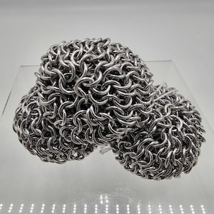 May include: A cluster of three interconnected spheres constructed from polished silver-colored metal rings. The individual rings are linked together in a dense, repeating chainmail pattern, creating a textured, spherical surface with a metallic sheen.