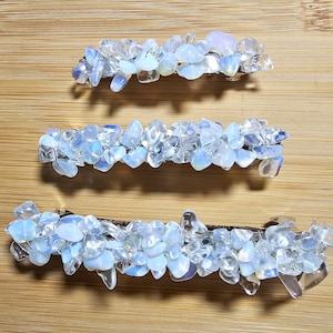 May include: Three hair barrettes, each decorated with iridescent, pale blue and clear crystal-like stones. The stones are arranged in a clustered pattern on a dark metal base, creating a shimmering effect. The barrettes vary slightly in size.