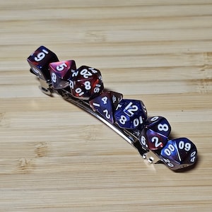 Puede incluir: Una pinza para el pelo plateada con seis dados iridiscentes unidos. Los dados son rojos y azules con números blancos. Los dados son un d20, un d12, un d10, un d8, un d6 y un d4.