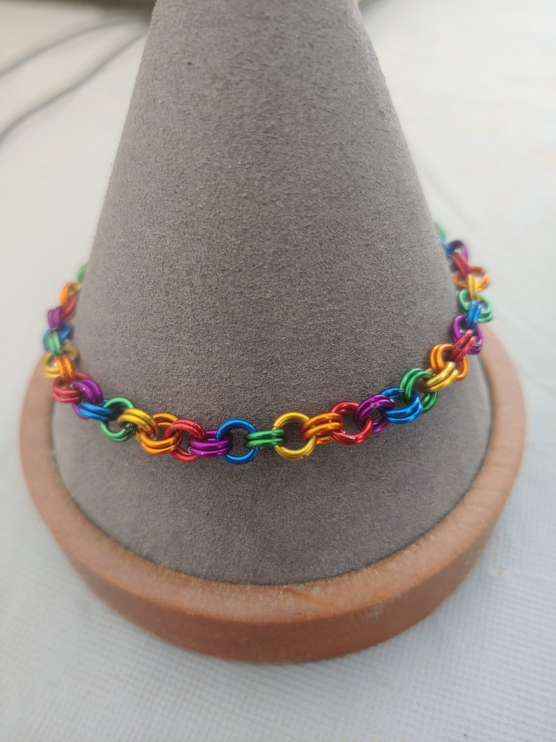 May include: A rainbow colored chain bracelet made of metal rings. The bracelet is displayed on a gray cone-shaped stand.