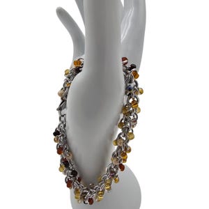 Puede incluir: Una pulsera de cadena plateada con cuentas colgantes en tonos ámbar, marrón y amarillo. La pulsera se exhibe en una forma de mano blanca, mostrando su intrincado diseño y variaciones de color.