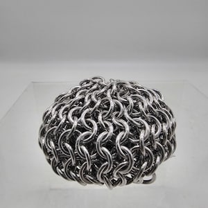 May include: A dense, spherical cluster of interconnected silver-colored metal rings forming a chainmail pattern. The individual circular links are tightly woven together, creating a textured, metallic surface with reflective highlights throughout.