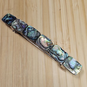 May include: A silver hair clip with a row of iridescent abalone shell squares. The shell has shades of blue, green, and purple.
