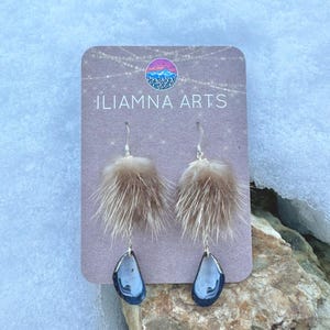 May include: A pair of dangle earrings with brown fur accents and teardrop-shaped blue and silver pendants. The earrings are displayed on a card with the text "ILIAMNA ARTS" and a mountain logo.