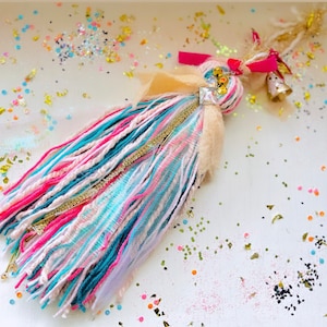 Peut inclure: Un pompon coloré avec du fil rose, bleu et blanc, accentué de paillettes dorées et d'un strass brillant. Le pompon est attaché à un ruban rose et un nœud en tissu beige.