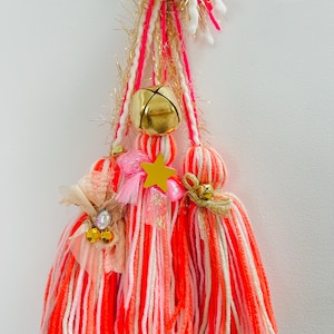 Könnte beinhalten: Handgefertigte Quasten in Orange-, Rosa- und Weißtönen, verziert mit goldenen Glocken, einem gelben Stern und dekorativen Verzierungen. Die Quasten sind aus Garn gefertigt und hängen vor einem weißen Hintergrund.