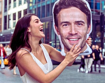 May include: A large, white pillow in the shape of a man's face. The pillow features a smiling man with brown hair and a white shirt. The background shows a city street with buildings and people.