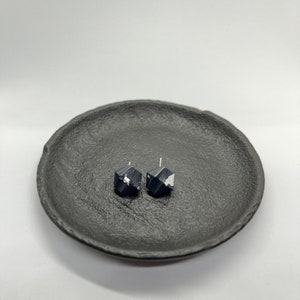 Könnte beinhalten: Ein Paar schwarze geometrische Ohrstecker mit silbernen Akzenten. Die Ohrringe werden auf einem strukturierten schwarzen Teller vor einem weißen Hintergrund präsentiert. Die Ohrringe haben ein facettiertes Design und sind aus dunklem Material.