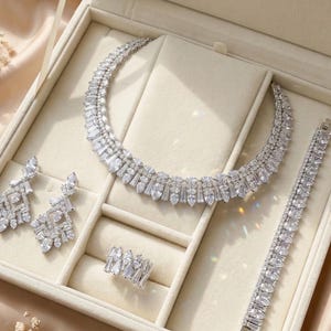 May include: A complete jewelry set displayed in a cream-colored box. The set includes a necklace, earrings, bracelet, and ring, all adorned with clear, sparkling stones. The jewelry is arranged on a cream-colored velvet-like surface.