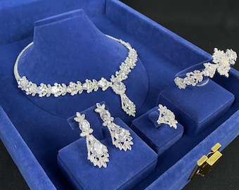 May include: A silver-toned bridal jewelry set displayed in a blue velvet box. The set includes a necklace, earrings, ring, and bracelet, all adorned with clear, sparkling gemstones. The necklace is displayed on a mannequin.