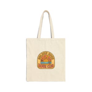 Peut inclure: Sac fourre-tout crème avec un graphisme coloré. Le graphisme présente une scène de coucher de soleil avec les mots "GOLDEN HOUR" et "HOUR CLUB" dans une police rétro. Le sac a deux poignées pour le transport.