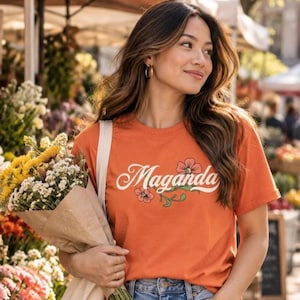 May include: An orange short-sleeved t-shirt featuring the word Maganda in white cursive script, accented with a small illustration of pink flowers and green leaves. The shirt is paired with blue denim jeans and a bouquet of flowers.