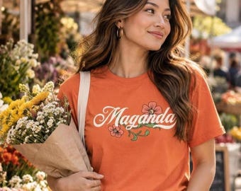 May include: An orange short-sleeved t-shirt featuring the word Maganda in white cursive script, accented with a small illustration of pink flowers and green leaves. The shirt is paired with blue denim jeans and a bouquet of flowers.