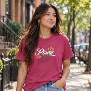 May include: A dark pink short-sleeved t-shirt featuring the word Pinay in white cursive script, accented by two orange flowers with green stems and leaves centered on the chest, paired with light blue denim jeans.
