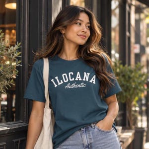 May include: A dark teal short-sleeve t-shirt featuring the white text ILOCANA in an arched block font with the word Authentic written in a white cursive script underneath. The shirt is paired with light blue denim jeans and a cream tote bag.