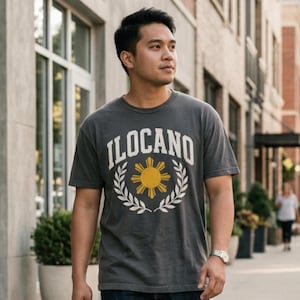 May include: A charcoal gray short-sleeve t-shirt featuring the text ILOCANO in a white arched font above a yellow sun icon and a white laurel wreath graphic centered on the chest. The shirt has a crew neckline and a standard fit.