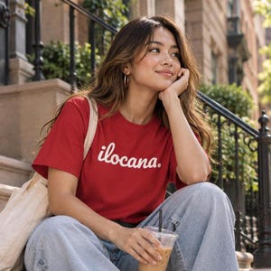 May include: A red short-sleeved t-shirt featuring the word ilocana. printed in white cursive script across the chest. The shirt is paired with light blue denim jeans and a beige canvas tote bag, creating a casual and everyday look.