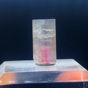 May include: A clear, faceted, pink and white tourmaline crystal with a rectangular shape. The crystal is sitting on a white display stand.