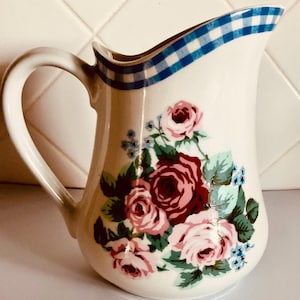May include: White ceramic pitcher with a curved handle and a blue and white checkered rim. The pitcher is decorated with a floral design featuring pink and red roses, green leaves, and small blue flowers.