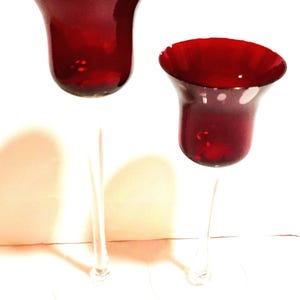 May include: Two tall candle holders with deep red glass bowls and clear glass stems. The bowls have a wide, flared rim and a rounded base. Each holder stands on a clear, circular glass base, with one holder being noticeably taller than the other.