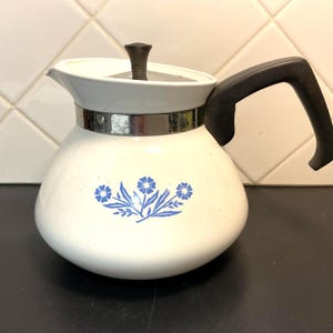 May include: White ceramic coffee pot with a black handle and lid. The pot features a silver band around the middle and a blue floral design. The pot is a classic kitchenware item.