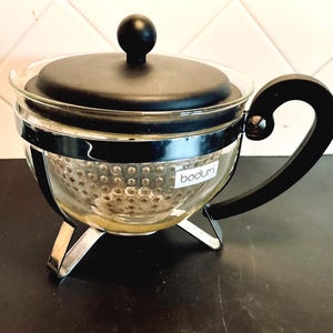 May include: A clear glass tea infuser with a black lid and handle. The infuser has a stainless steel band and sits on three silver legs. The brand name "bodum" is visible on the glass.