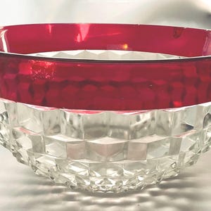 May include: A round glass bowl featuring a clear, faceted geometric pattern on the lower portion and a smooth, deep red band around the upper rim. The bowl has a wide opening and a curved base, showcasing a contrast between the transparent crystal-like texture and the solid, vibrant red glass color at the top.