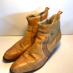 May include: A pair of tan leather ankle boots. The boots have a pointed toe, a side zipper, and a woven leather detail on the side. The boots have a pull tab at the top and a low heel. The leather has a worn appearance.