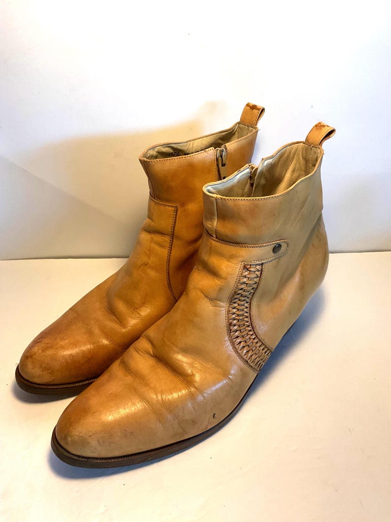 May include: A pair of tan leather ankle boots. The boots have a pointed toe, a side zipper, and a woven leather detail on the side. The boots have a pull tab at the top and a low heel. The leather has a worn appearance.