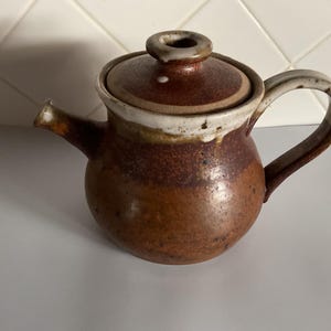 May include: A handcrafted ceramic teapot with a lid. The teapot is primarily brown with a white rim and handle. The spout is orange. The teapot is designed for brewing tea and has a rustic, handmade aesthetic.