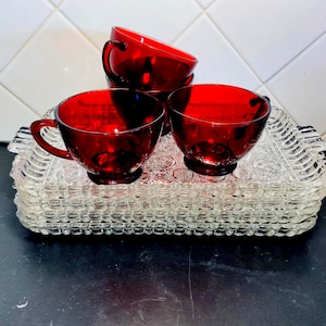 May include: A stack of clear glass serving trays with a beaded edge, topped with four ruby red glass teacups. The cups have handles and sit on the trays. The background is a white tiled wall.