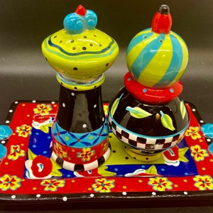 May include: A set of two colorful ceramic salt and pepper shakers with a black and white checkered design. The shakers are sitting on a tray with a red, white, and blue floral pattern.