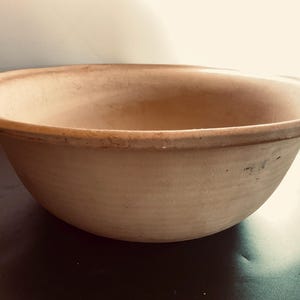May include: A large, round, beige ceramic bowl with a wide, flat rim. The bowl features a smooth, matte finish with subtle variations in color across its surface, showing a simple, functional design with a deep basin and a thick, rounded edge.