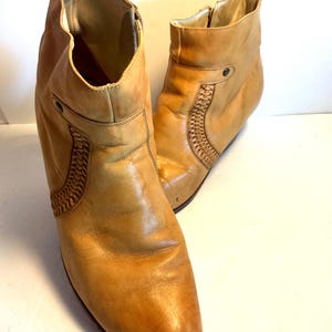 May include: A pair of tan leather ankle boots. The boots have a pointed toe, a small heel, and decorative woven leather detailing on the sides. The boots have a zipper closure and a pull tab at the top of the boot.