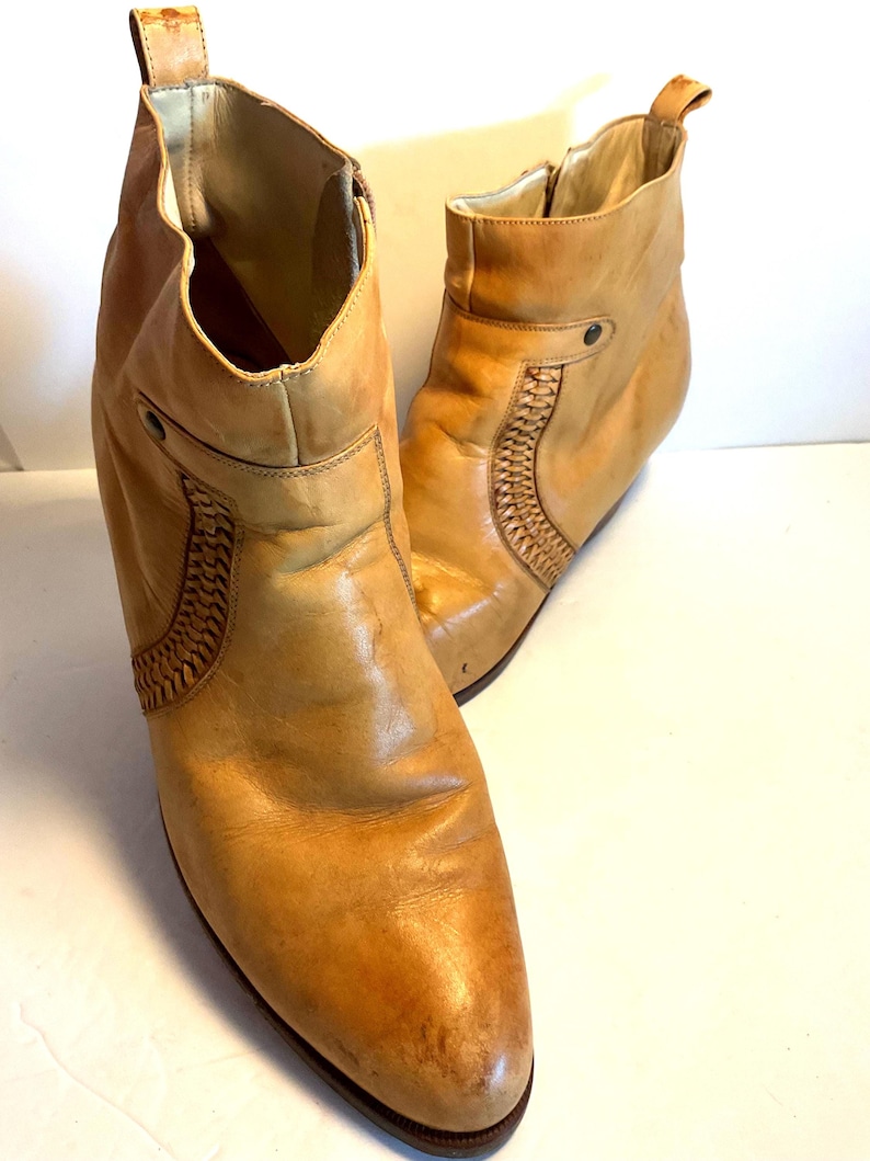 May include: A pair of tan leather ankle boots. The boots have a pointed toe, a small heel, and decorative woven leather detailing on the sides. The boots have a zipper closure and a pull tab at the top of the boot.