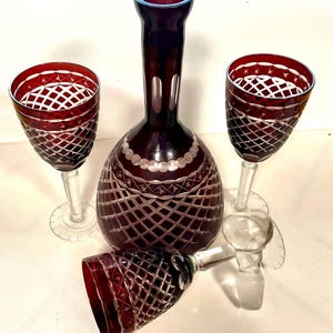 May include: A set of deep red cut-to-clear glass consisting of a decanter with a matching stopper and three stemmed goblets. Each piece features a diamond-cut lattice pattern with star motifs, clear glass stems, and circular bases.