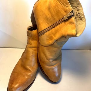 May include: A pair of tan leather ankle boots with a zipper closure. The boots have a worn appearance, with visible creases and wear marks. The interior of one boot displays printed text.
