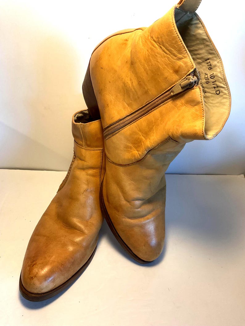 May include: A pair of tan leather ankle boots with a zipper closure. The boots have a worn appearance, with visible creases and wear marks. The interior of one boot displays printed text.