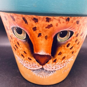 May include: A hand-painted terracotta flower pot with a blue rim and a cheetah face painted on the front. The cheetah has brown spots and a white nose.