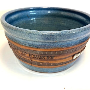May include: A handmade ceramic bowl with a blue glazed interior and rim. The exterior features a band of brown and tan with the text "WHAT SHALL SEPARATE US? ROMANS 8:31". A white decorative element is also present.