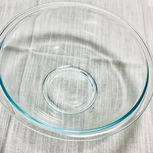 May include: A clear glass mixing bowl with a light blue rim and a smaller, matching bowl inside. The bowl is sitting on a white and gray patterned surface. The bowl is empty.