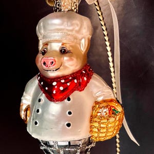 May include: A glass ornament shaped like a pig dressed as a chef, wearing a white hat, a white jacket with black buttons, a red polka-dot neckerchief, and black and white checkered pants, holding a gold-colored basket with a white ribbon hanger.