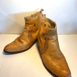 May include: A pair of tan leather ankle boots. The boots have a side zipper, a decorative woven panel, and a pull tab at the top. The soles are dark brown. The boots are likely vintage or pre-owned.