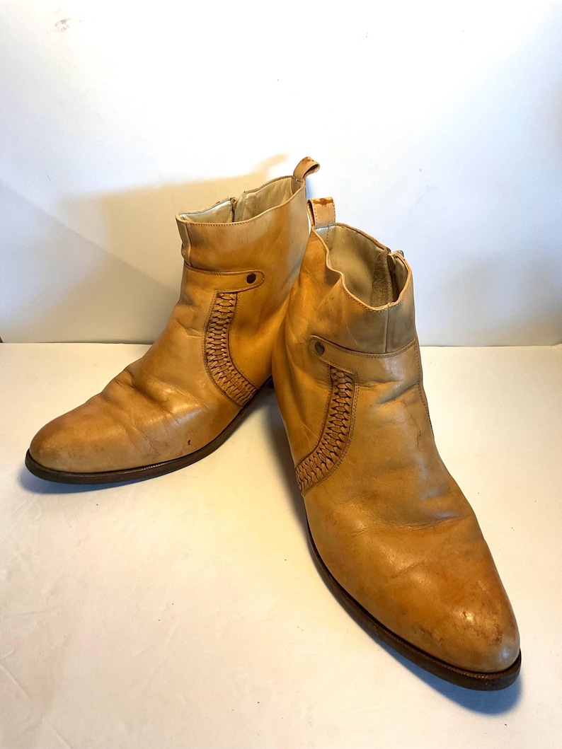 May include: A pair of tan leather ankle boots. The boots have a side zipper, a decorative woven panel, and a pull tab at the top. The soles are dark brown. The boots are likely vintage or pre-owned.