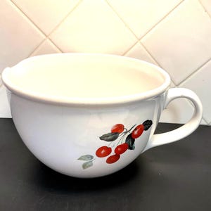 May include: White ceramic mixing bowl with a handle and a cherry branch design. The bowl has a wide opening and a curved shape. The cherry branch features red cherries and green leaves. The bowl is on a black surface.