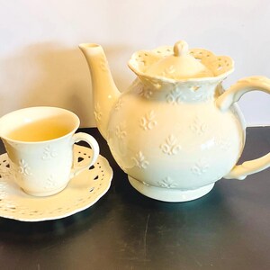 May include: A cream-colored ceramic teapot and a matching teacup on a saucer. The teapot features a lid with a scalloped, pierced edge and a repeating embossed floral pattern. The teacup and saucer also display the same embossed floral design.