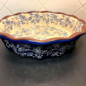 May include: Oval ceramic baking dish with a scalloped edge. The dish features a cream-colored interior with blue floral patterns. The exterior is navy blue with white floral designs, and the rim is brown. A decorative and functional kitchenware item.