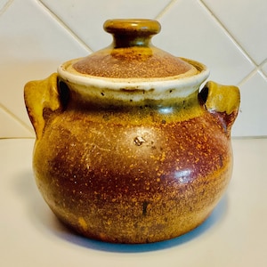 May include: A ceramic lidded pot with a rustic, speckled appearance. The pot is primarily brown with yellow and white accents, featuring two side handles and a rounded lid with a knob. This handcrafted piece is suitable for kitchen decor.