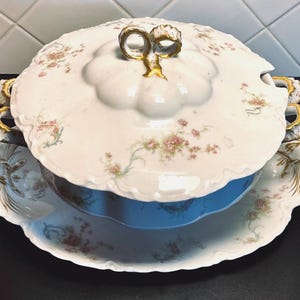 May include: A white porcelain serving dish with a blue interior, floral accents, and gold trim. The lid features a gold bow-shaped handle. The dish sits on a matching scalloped tray, perfect for serving soups or stews.